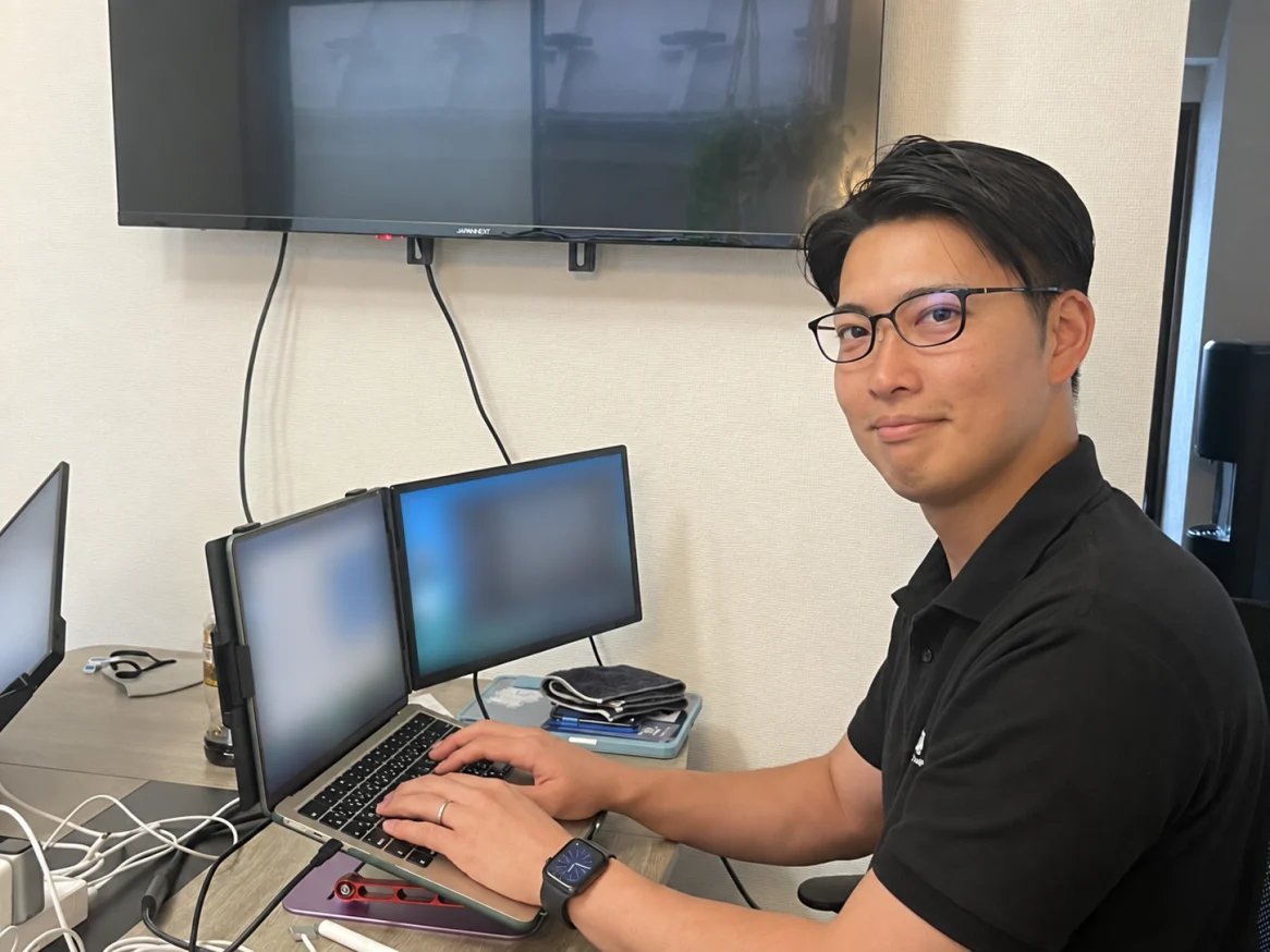 デスクでパソコンに手を置き、こちらに顔を向ける三瓶峻輔